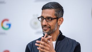 A telephoto shot of Sundar Pichai, clapping his hands at an event. In the background the Google logo is repeated on a wall, out of focus.