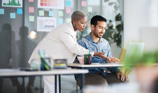 A business woman looking over a colleague's shoulder at some work he is doing and offering advice