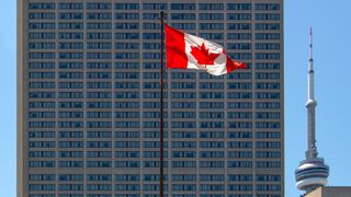 A Canadian flag, flying on a tall pole in front of a skyscraper.