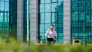 A man sat on a bench on the phone, with a laptop on his lap and a coffee sat next to him. It is a sunny day and he's sat in what looks like a business park, partly obscured by shrubs in the foreground, which are out of focus. It's ambiguous whether or not the man is working remotely.