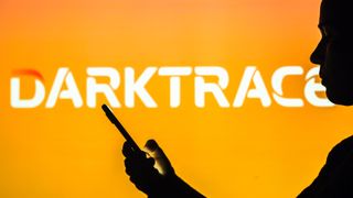 Darktrace logo pictured in the background of a silhouetted woman holding a mobile phone.