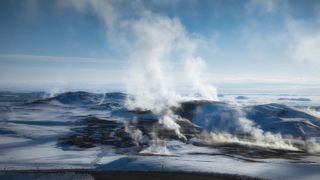 An aerial shot of the Krafla volcanic caldera in Iceland, which is used for geothermal green energy. Steam rises from the fissure in the earth, with pale blue water contrasted against the dark rocks.