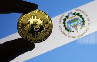 Fingers holding a 'bitcoin' in front of the flag for El Salvador