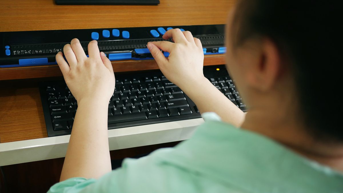 A close-up shot of a vision impaired person using a computer keyboard and braille display, to represent accessibility in tech. The person is shot from behind and is wearing smart casual clothing.